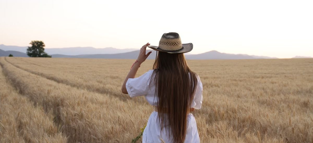 A woman stands in a wheat field adjusting her straw hat, back turned, during a serene sunset.