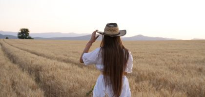 A woman stands in a wheat field adjusting her straw hat, back turned, during a serene sunset.