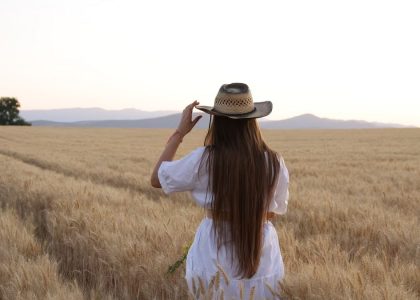 A woman stands in a wheat field adjusting her straw hat, back turned, during a serene sunset.