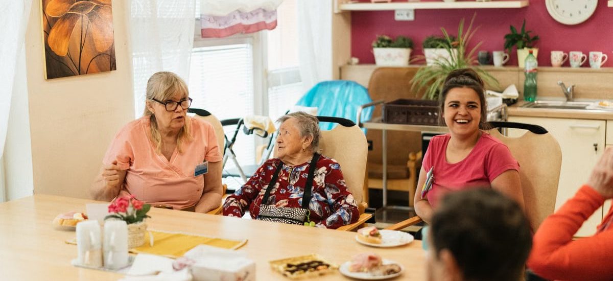 Elderly residents and caregivers enjoying a meal and conversation in a cheerful dining space.